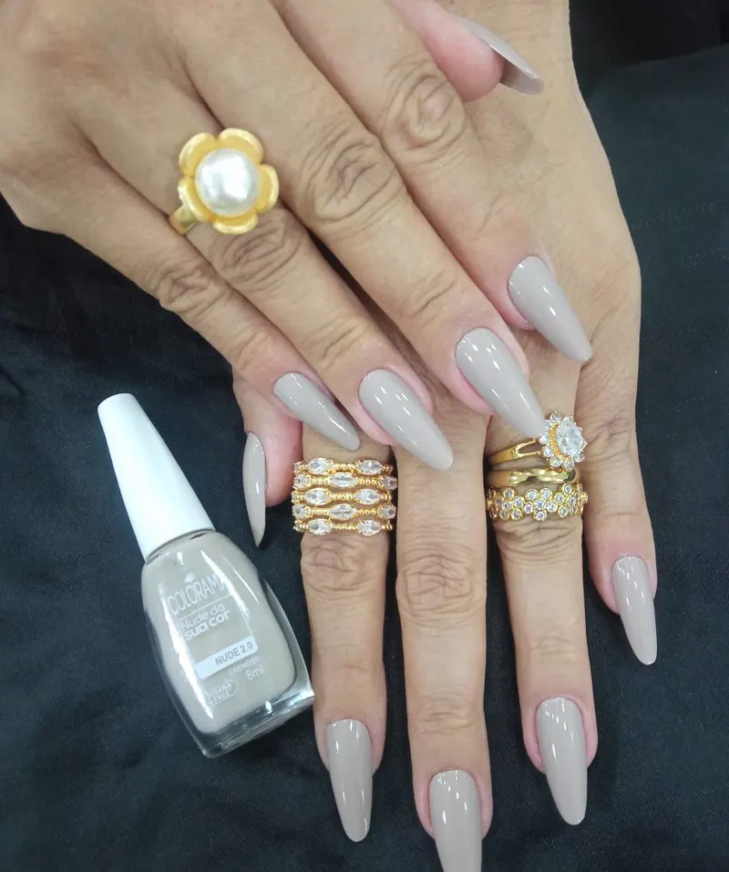 Mãos com unhas longas pintadas em tom nude, ao lado do esmalte usado